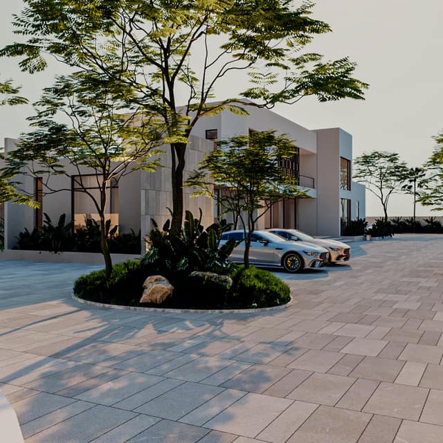 A broad forecourt in pale paving before the villa, centred on a round planted island with a tree whose shadow falls across the paving, two cars parked beside it, and planting held to the island and the forecourt's edges.